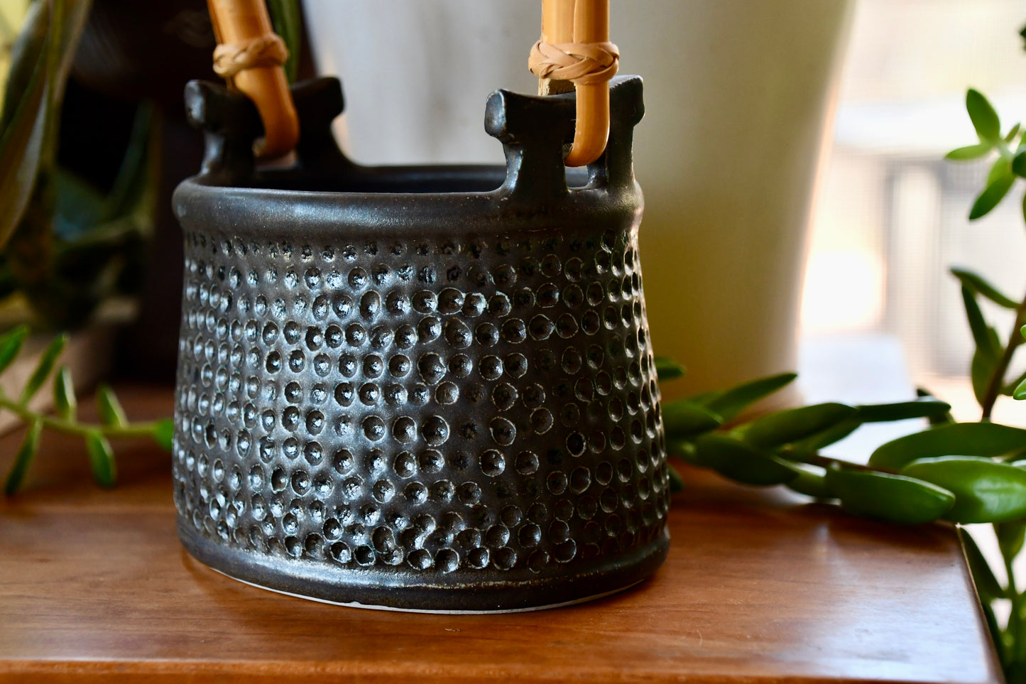 Black textured ceramic pot with bamboo handles on a wooden surface with plants in the background