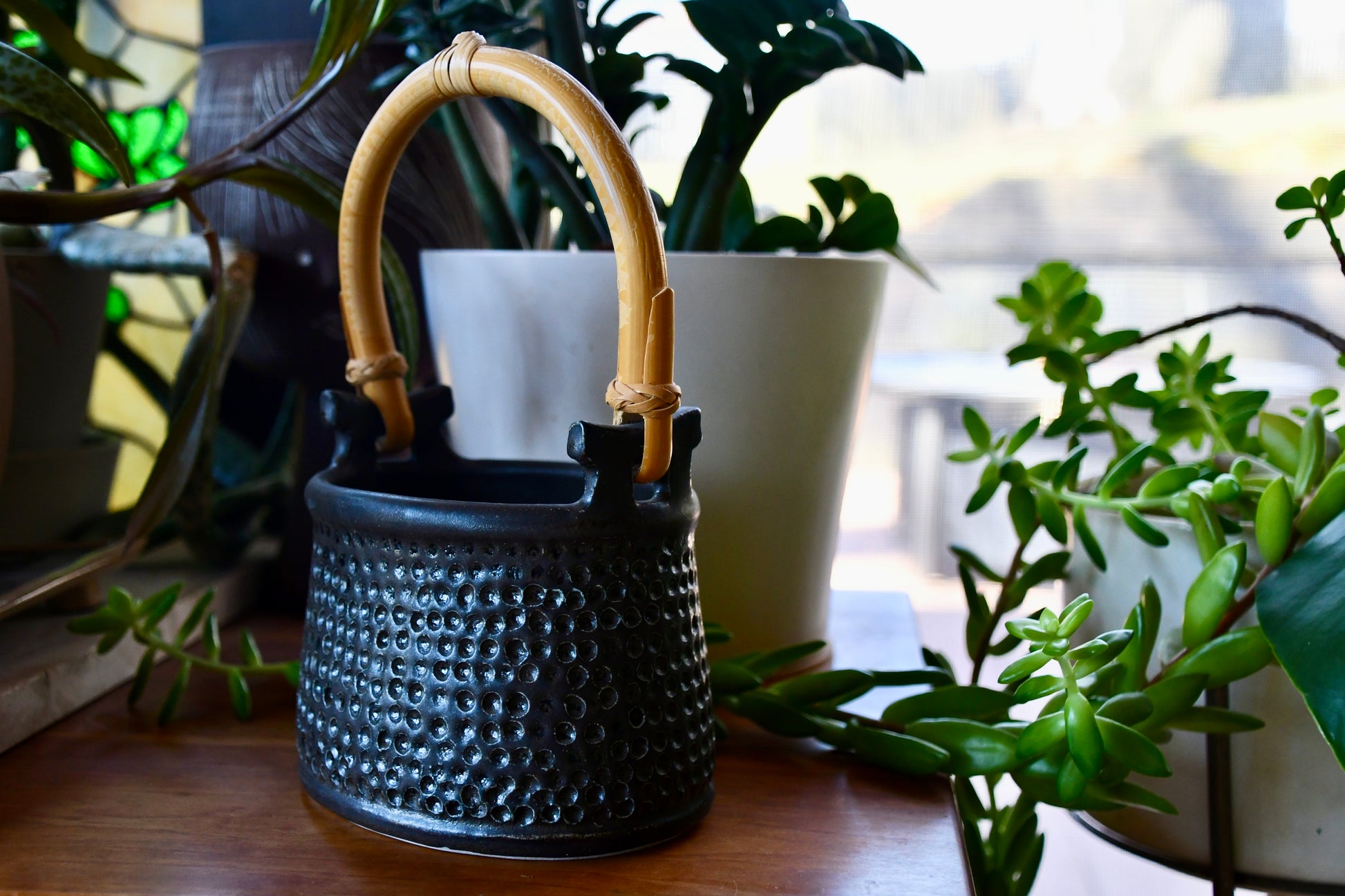 Black textured vase with bamboo handle on a wooden surface surrounded by plants