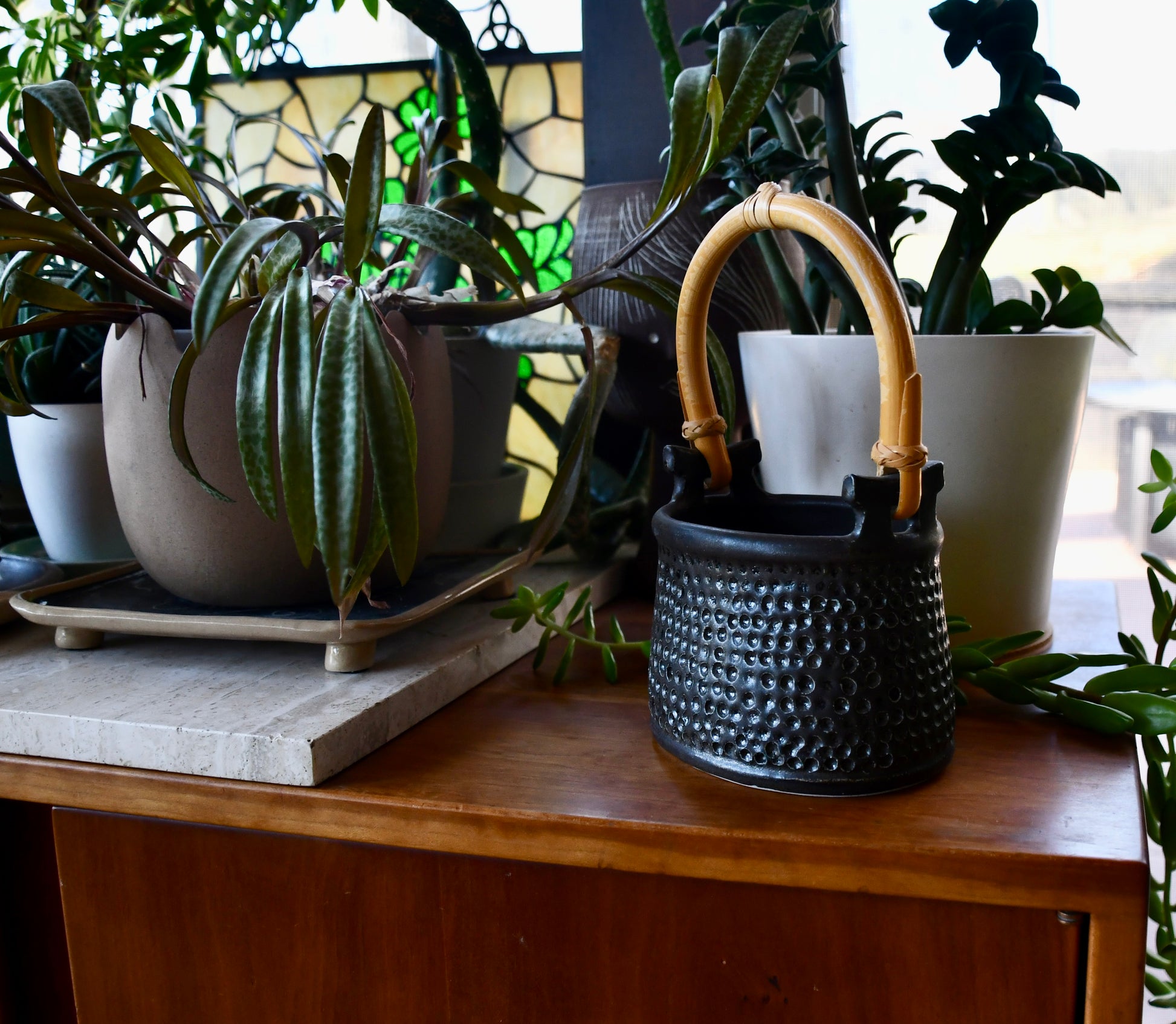 Black textured pot with a bamboo handle on a wooden surface surrounded by potted plants.