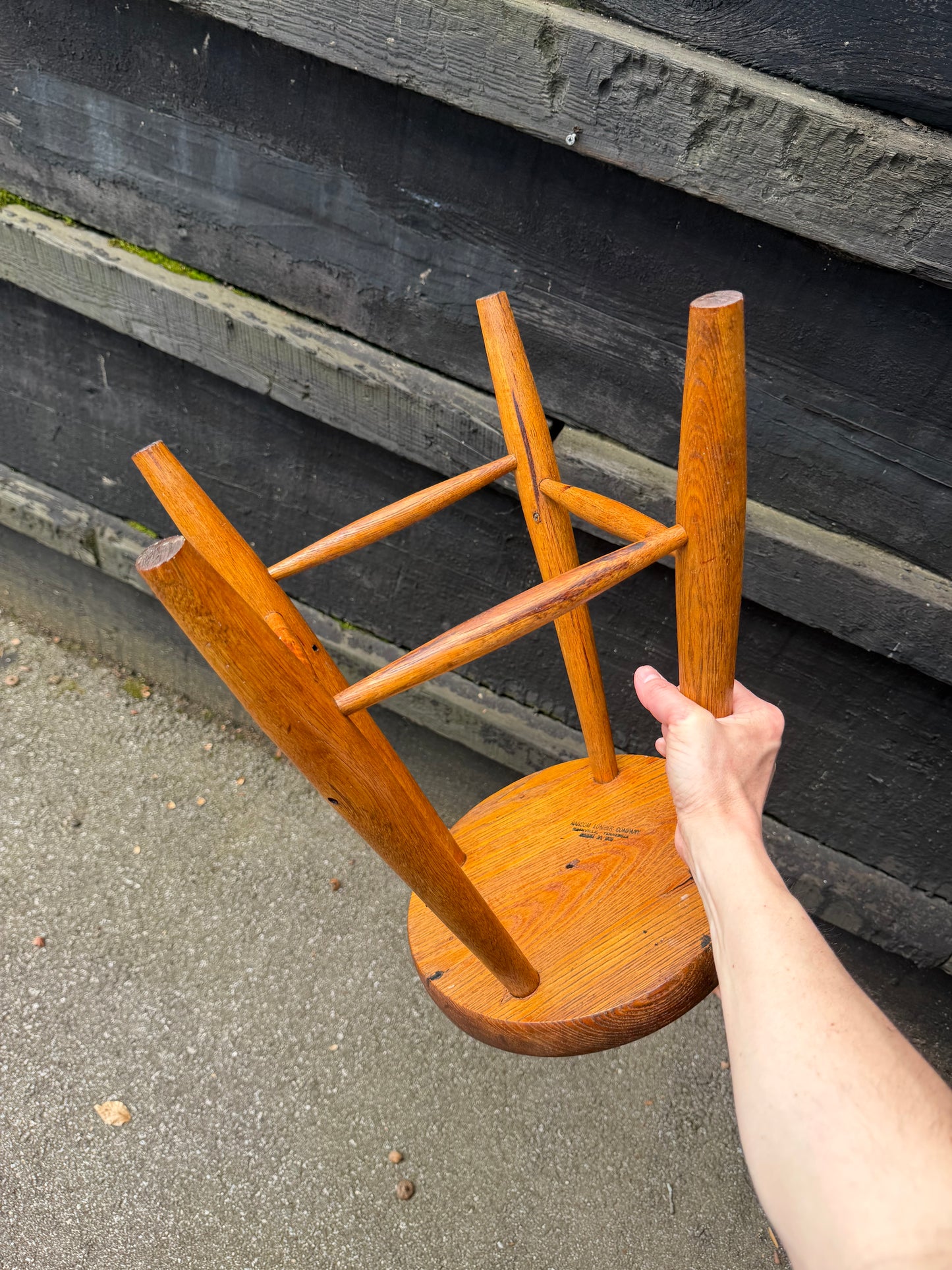 Wooden stool being held by a person on a concrete surface with a tire in the background