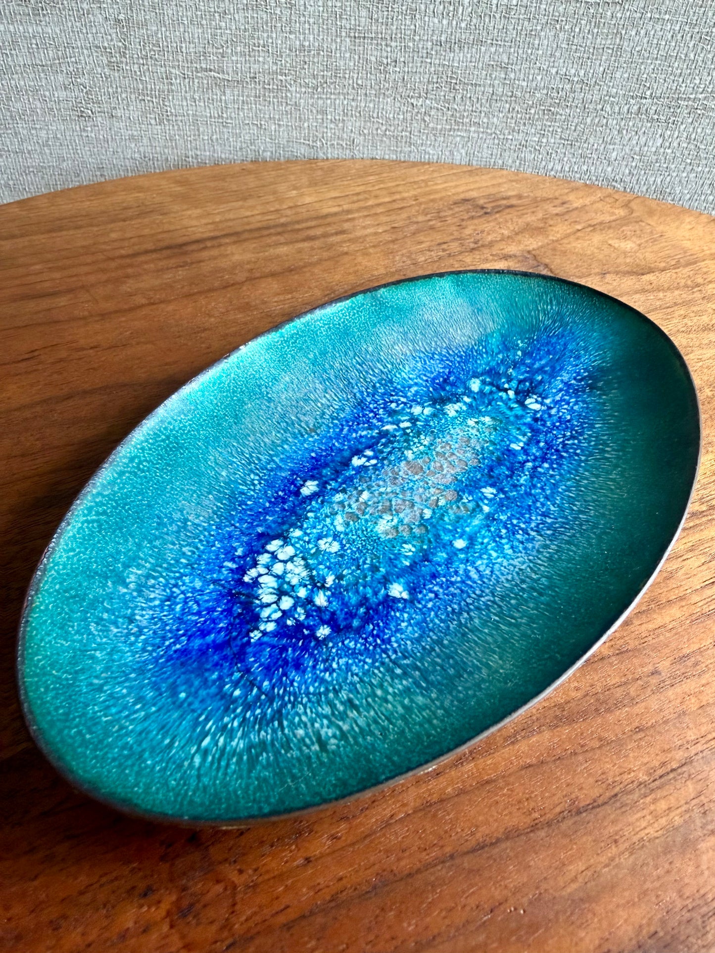 Oval-shaped enamel copper piece with blue and green gradient on a wooden surface