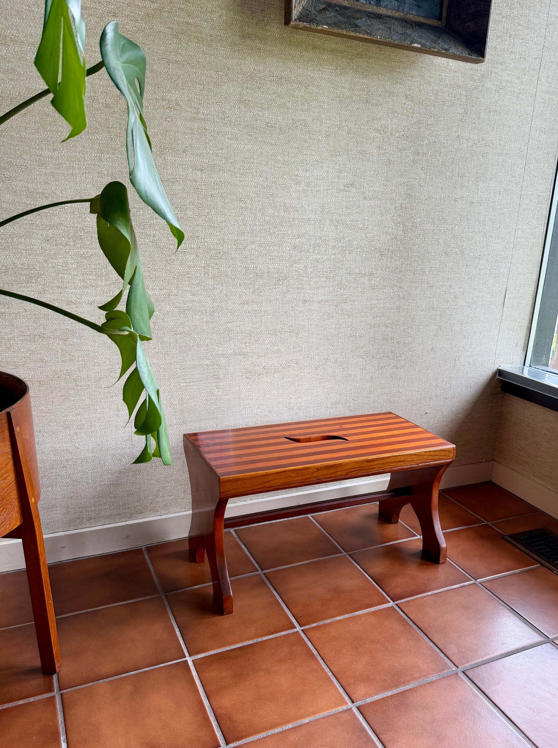 Wooden stool against a textured wall with a plant in the foreground.