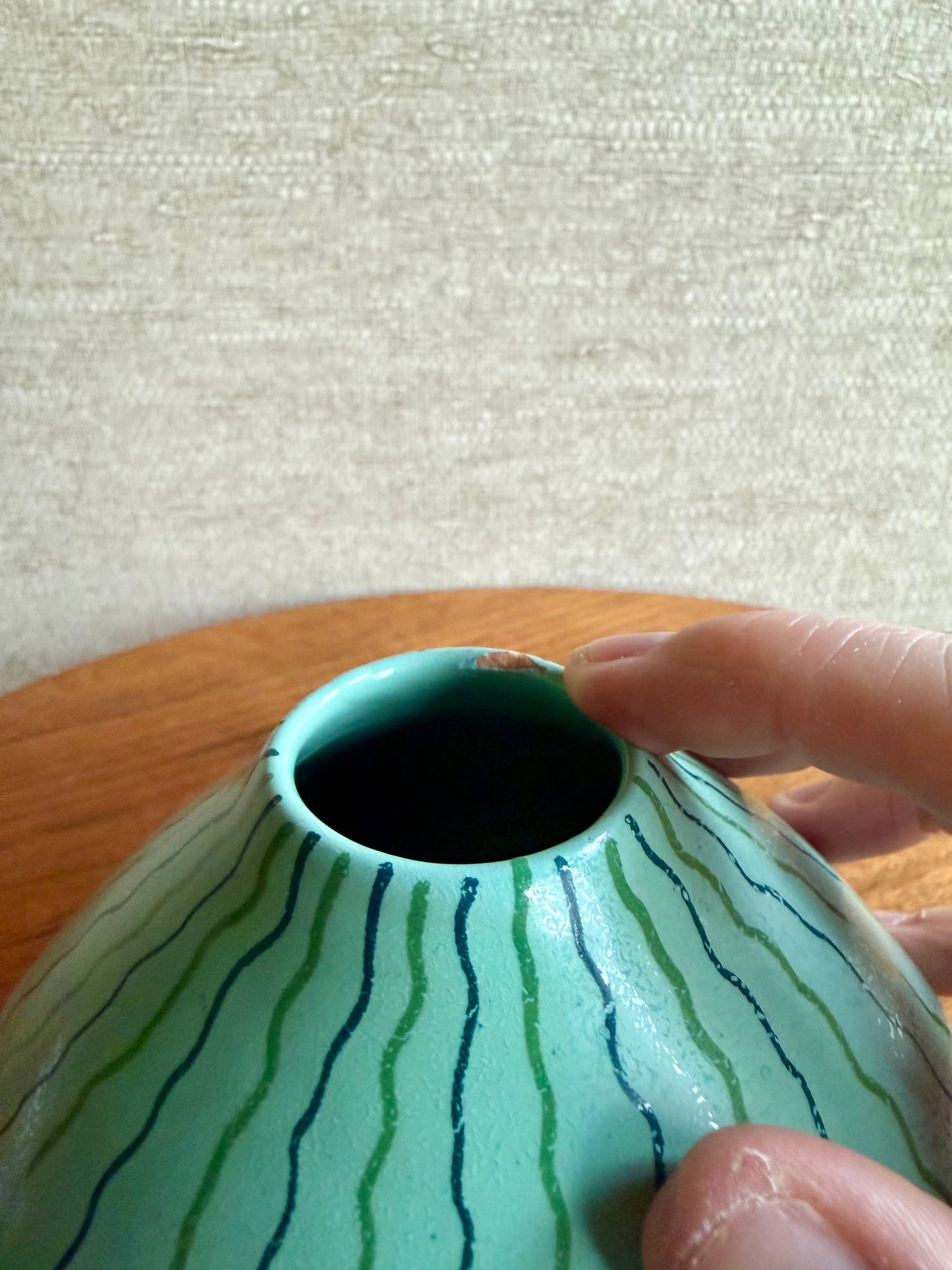 Hand holding a small green ceramic vase with wavy blue lines on a wooden surface.