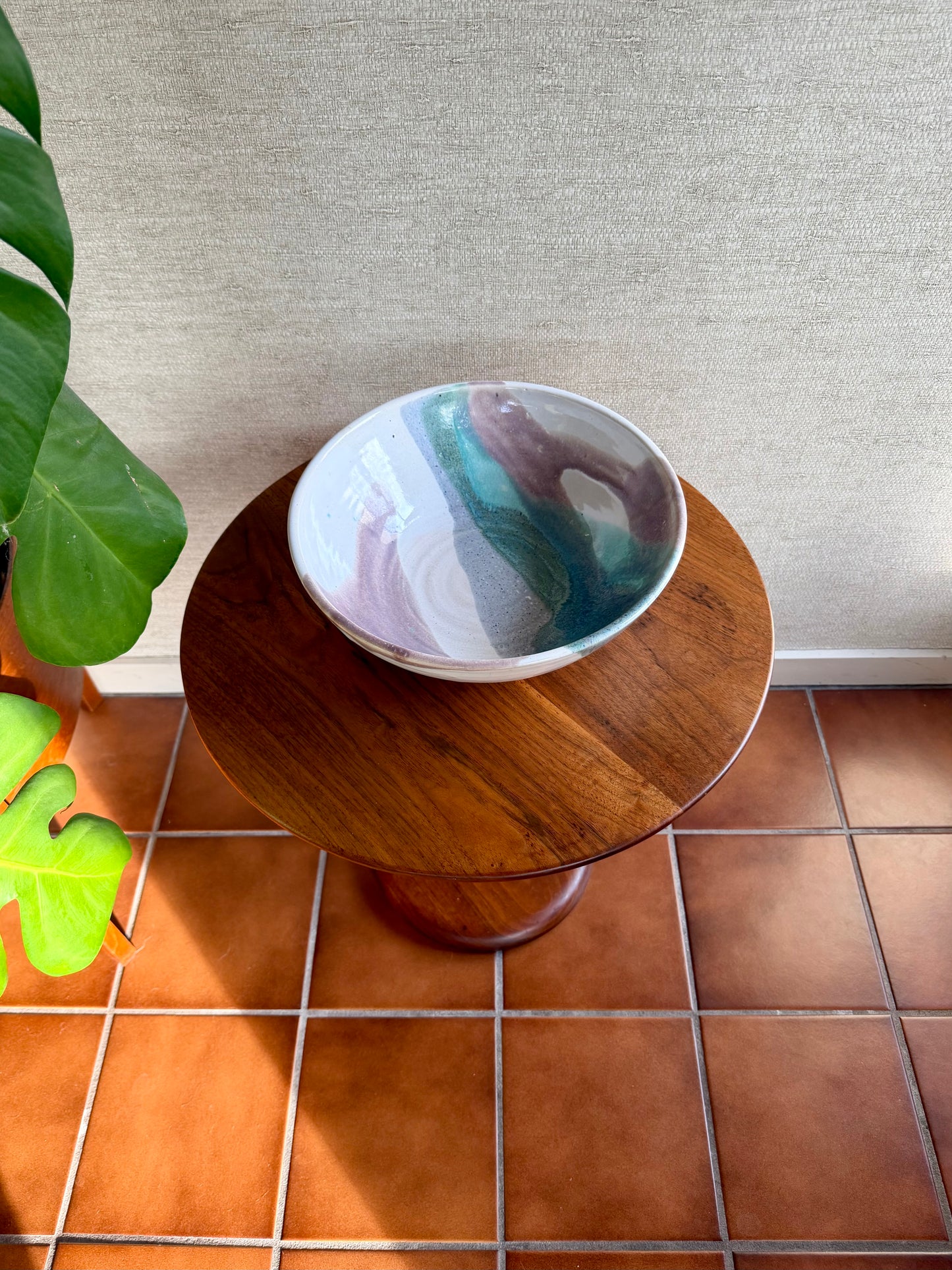 Small round wooden table with a ceramic bowl on a tiled floor next to a plant.