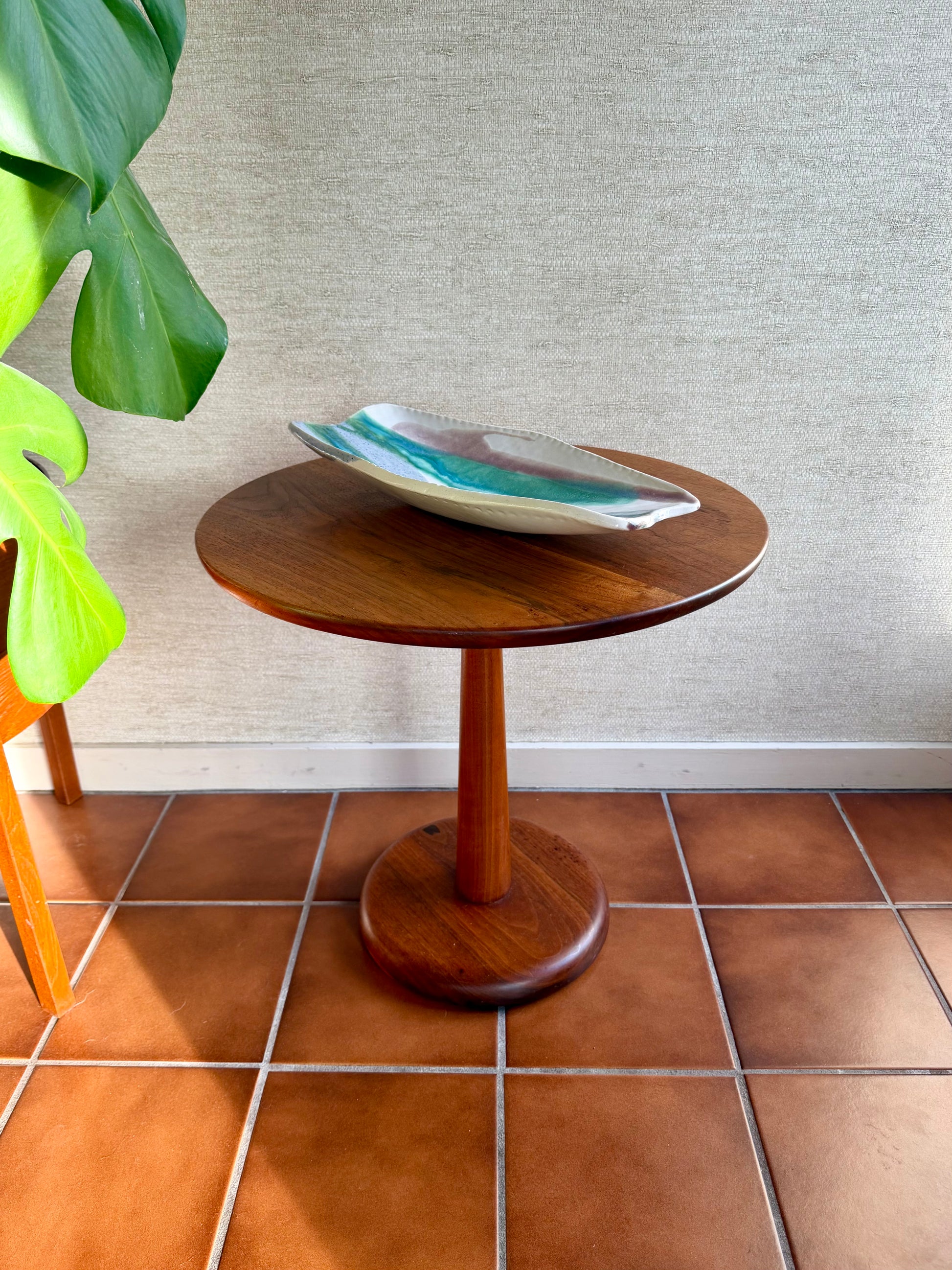 Wooden side table with a decorative bowl on a tiled floor next to a plant.