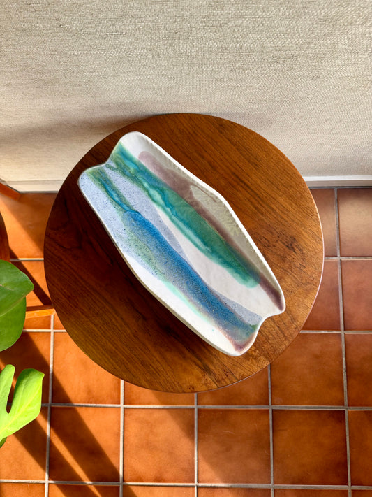 Ceramic dish with blue and green stripes on a wooden stand against a tiled wall.