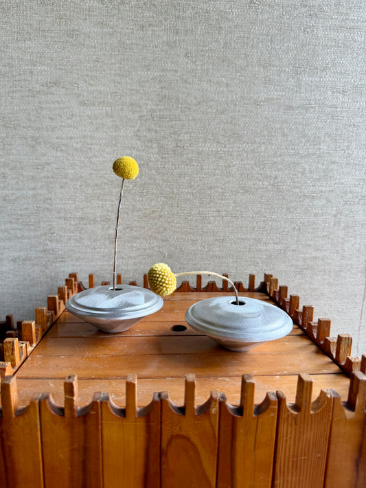 Two metal vases on a wooden stand with decorative elements against a textured wall.