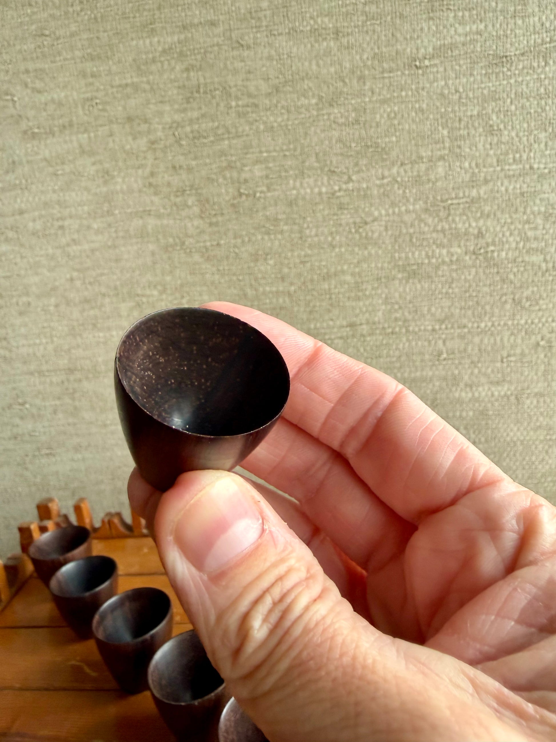 Hand holding a small black cup against a textured beige wall with more cups on a wooden surface.