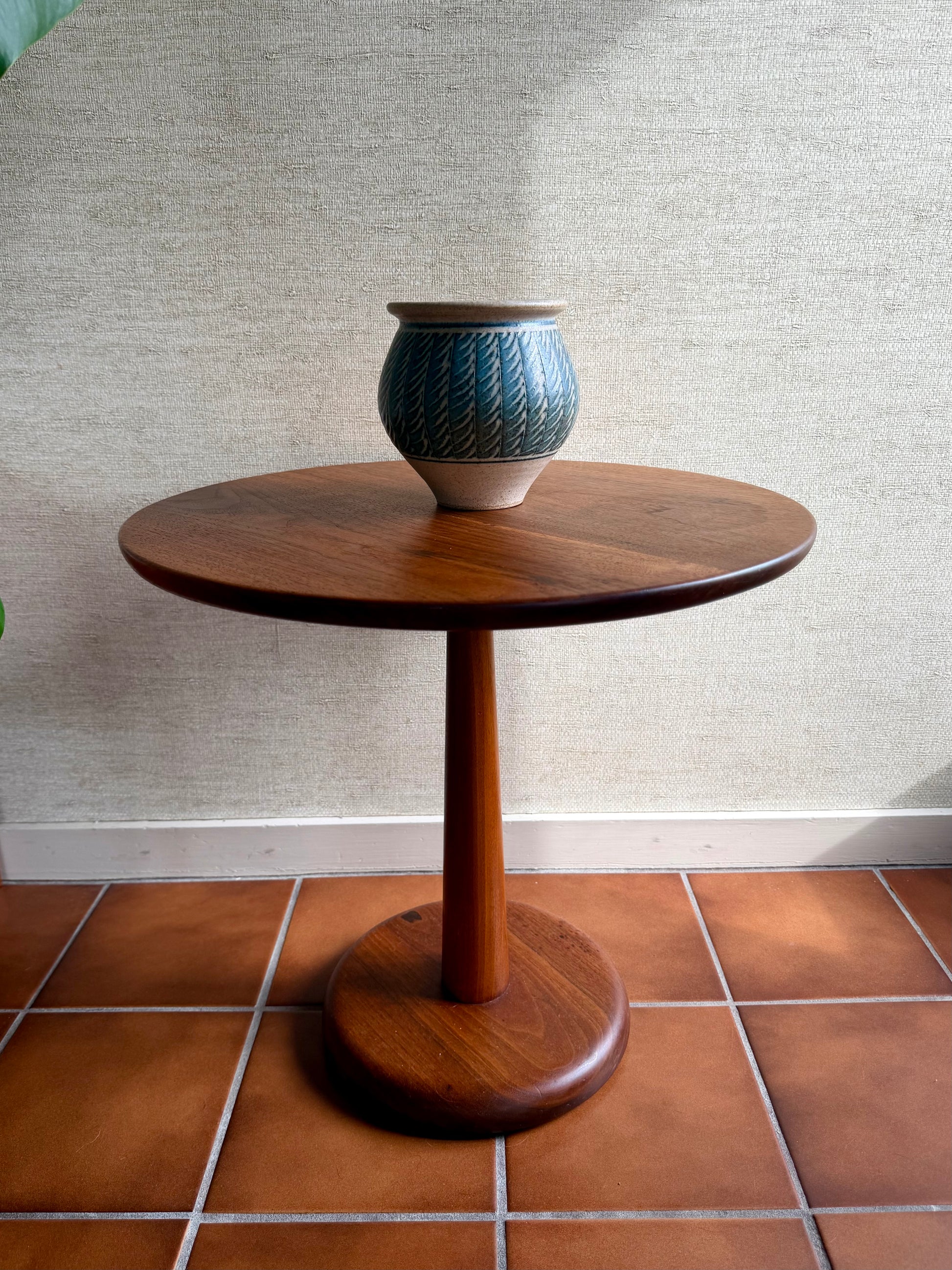Wooden side table with a ceramic vase on a tiled floor against a textured wall.