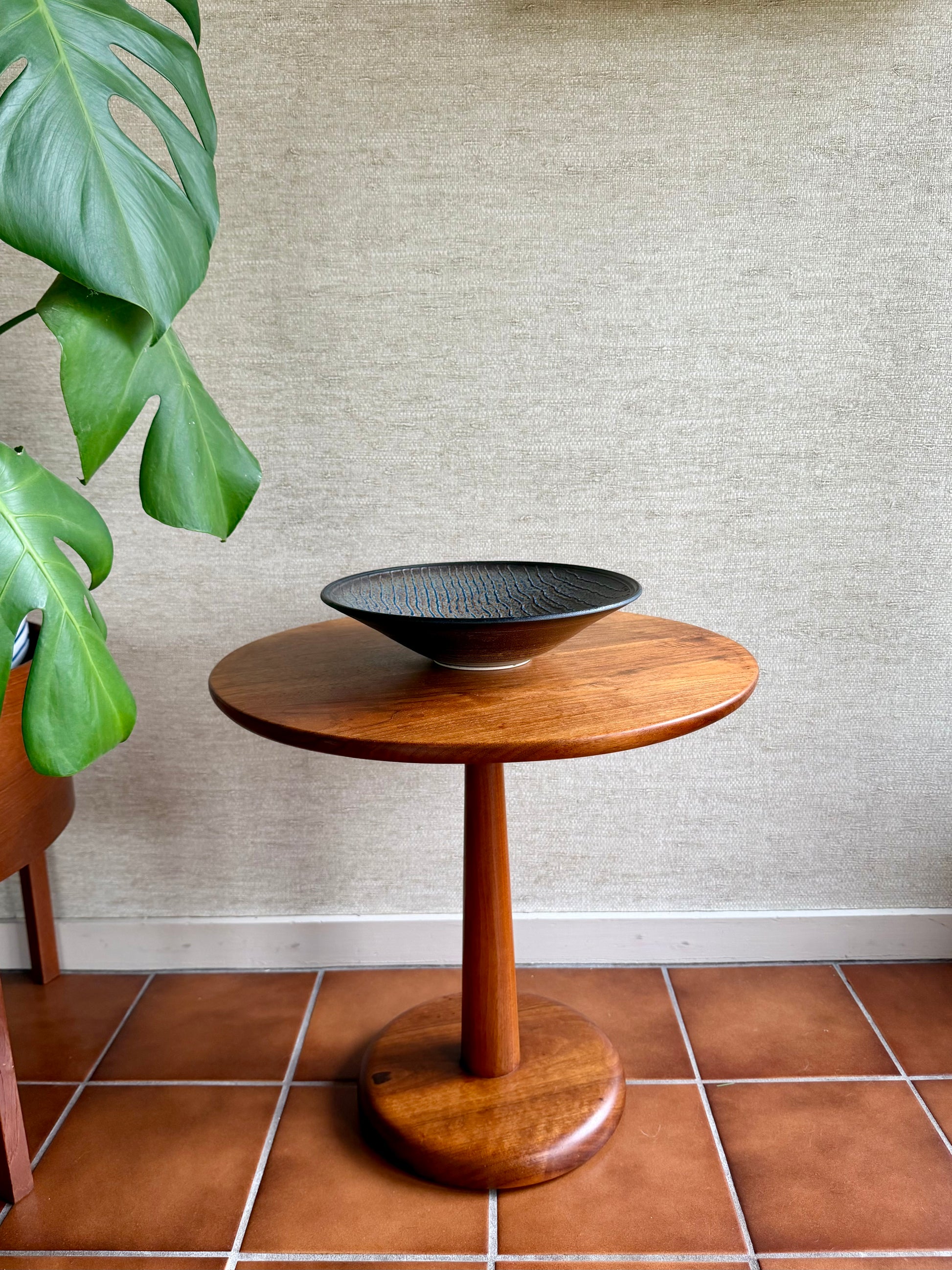 Wooden side table with a black bowl on a tiled floor next to a plant.