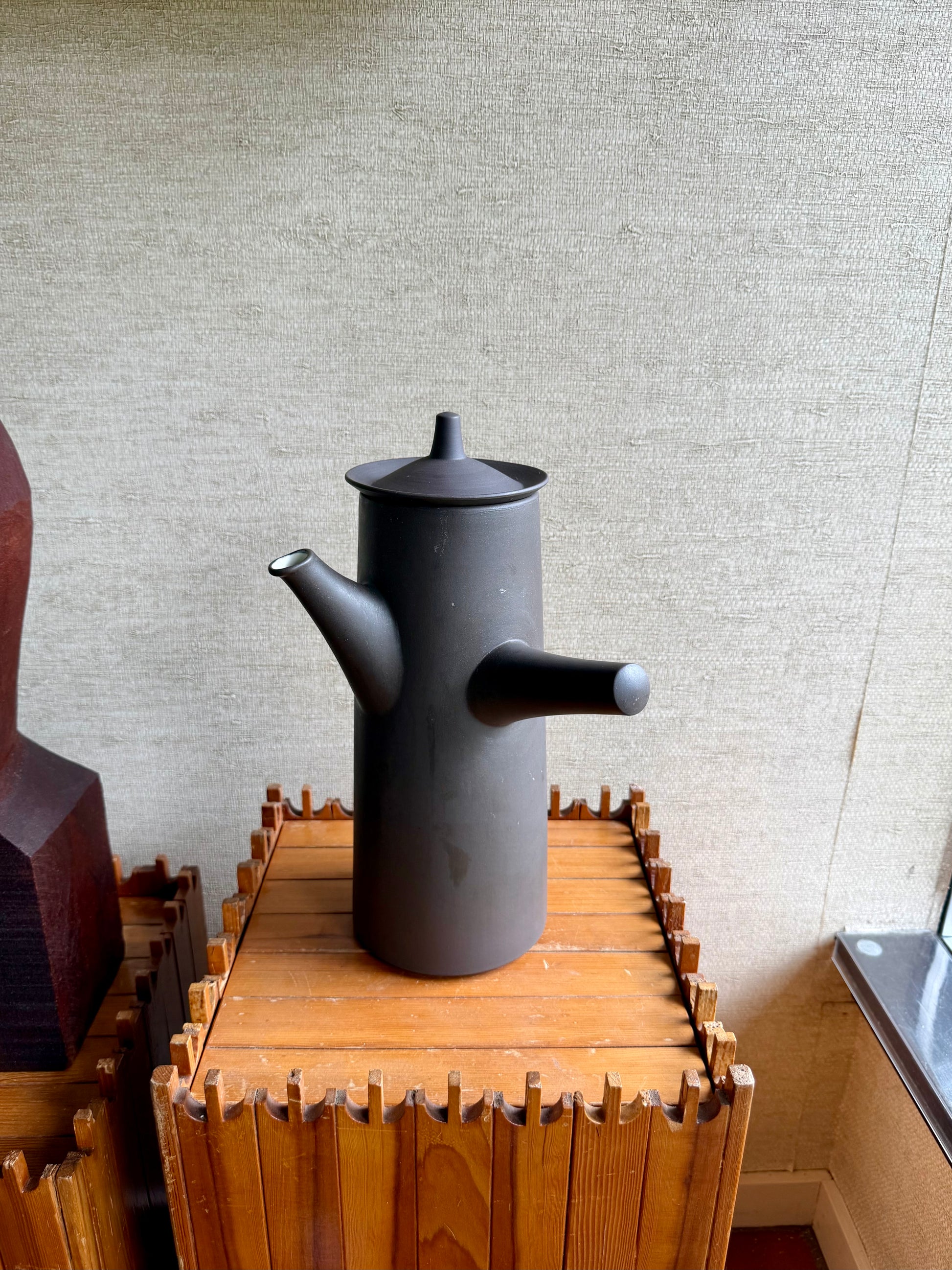 Black ceramic teapot on a wooden stand against a textured wall.