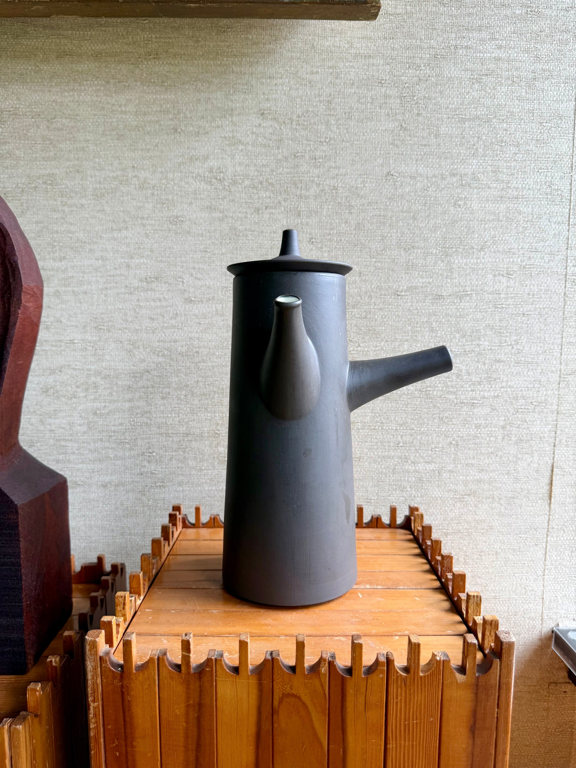 Black ceramic teapot on a wooden stand against a textured wall.