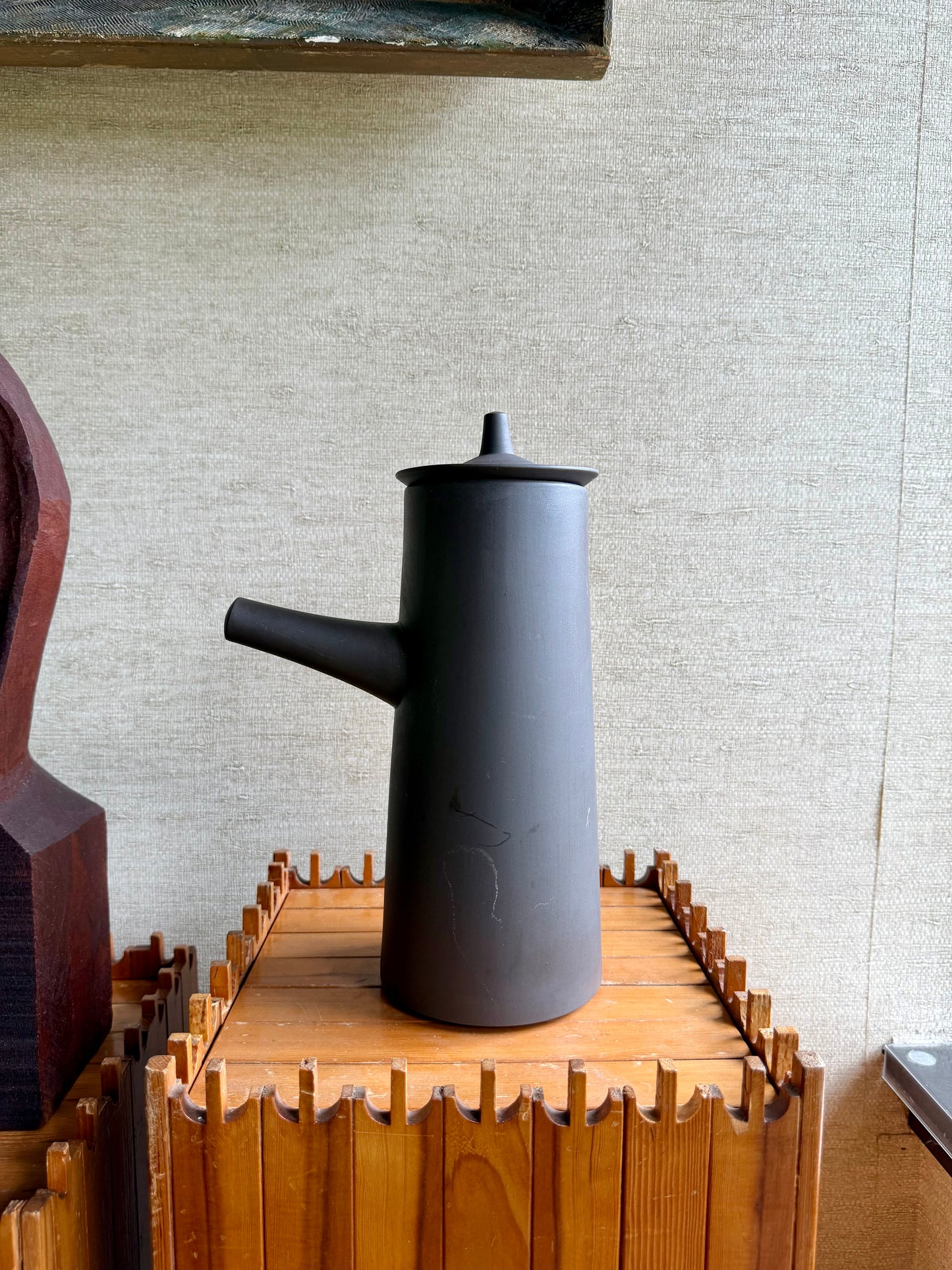 Black ceramic teapot on a wooden stand against a textured wall.