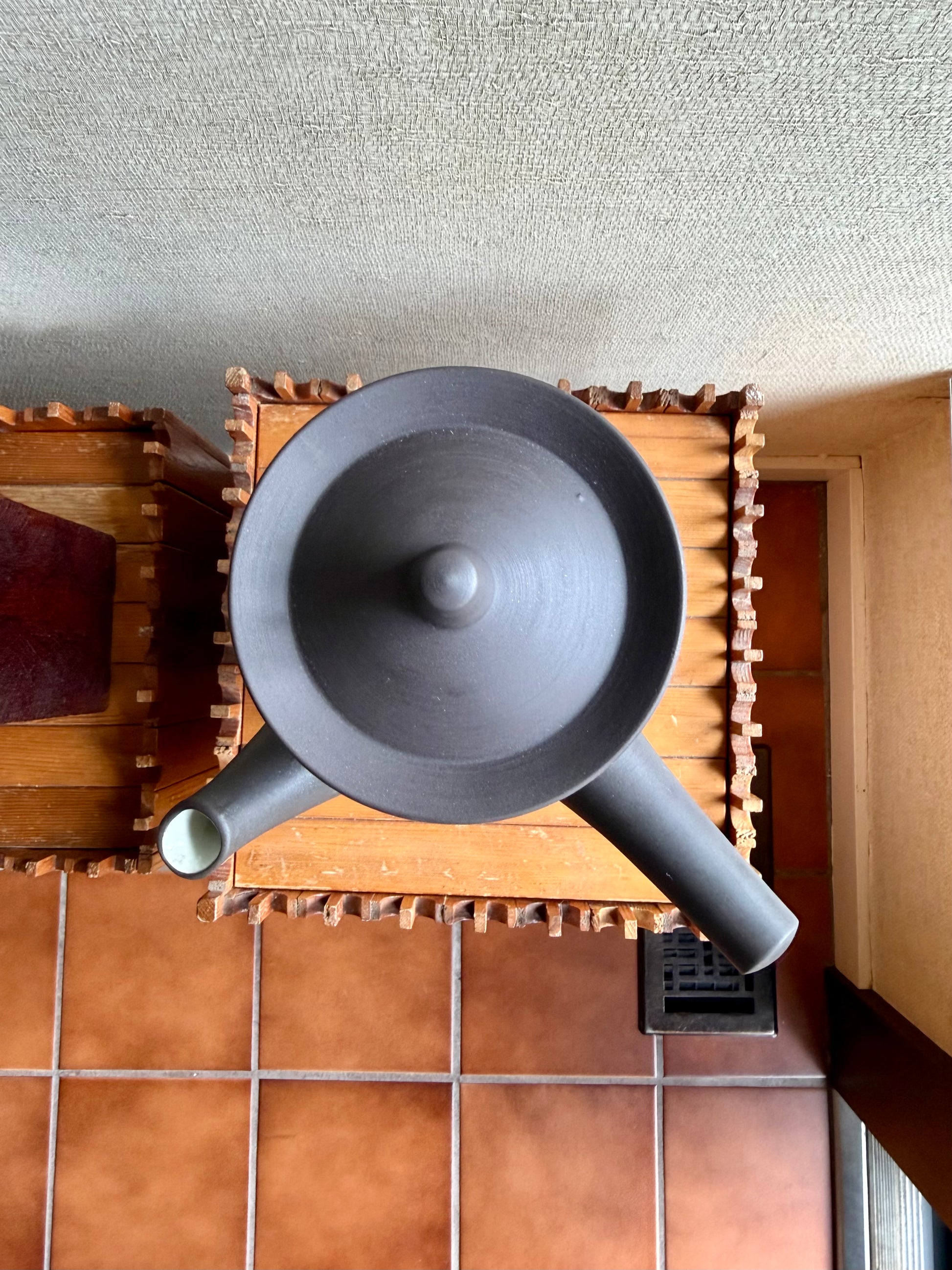 Black ceramic teapot on a wooden stand with a textured wall and tiled floor in the background