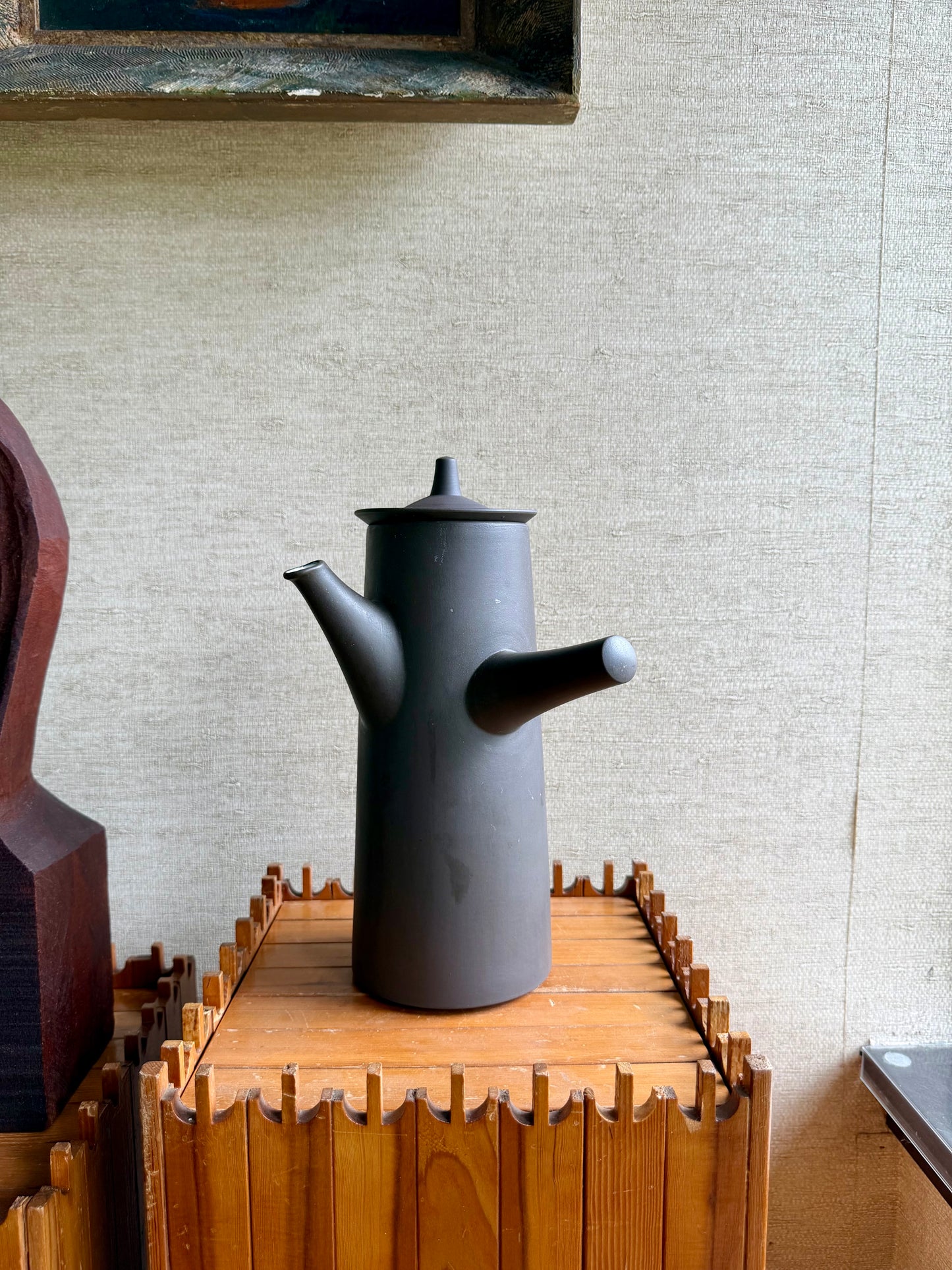 Black ceramic teapot on a wooden stand against a textured wall.