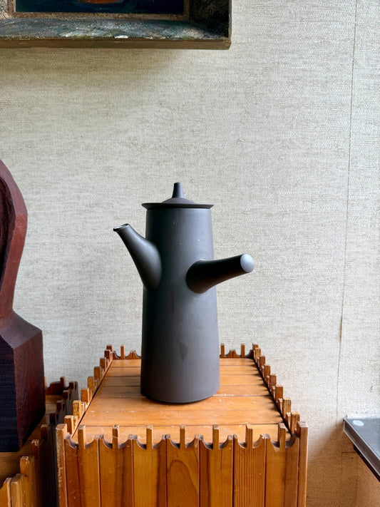 Black ceramic teapot on a wooden stand against a textured wall.