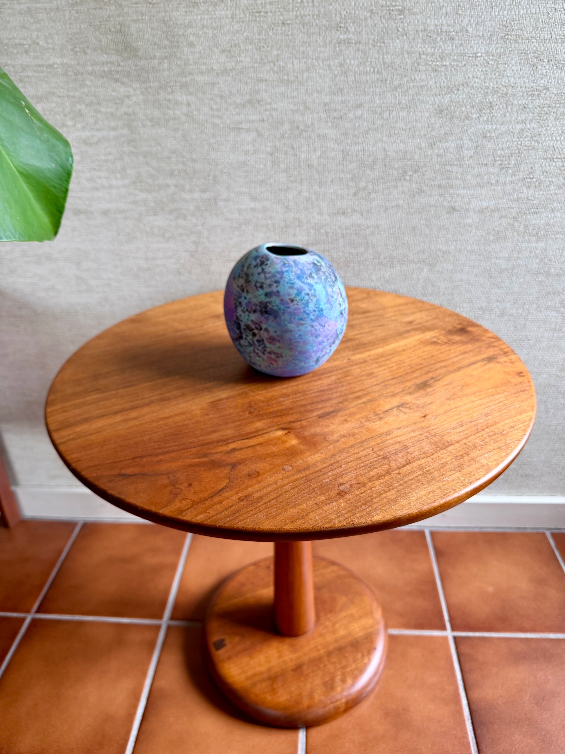 Wooden side table with a colorful vase on a tiled floor against a gray wall.