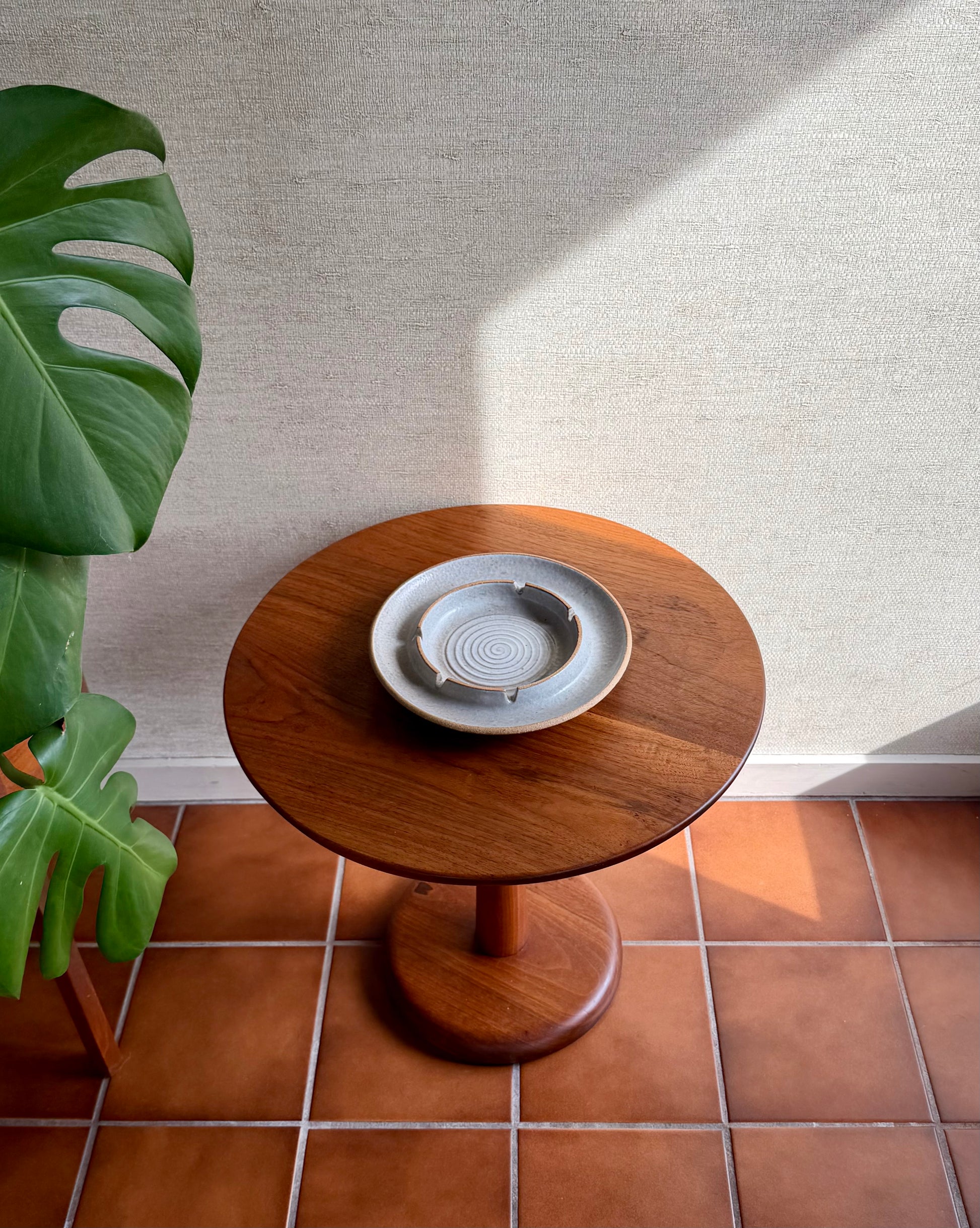 Wooden side table with a ceramic plate on a tiled floor next to a plant.