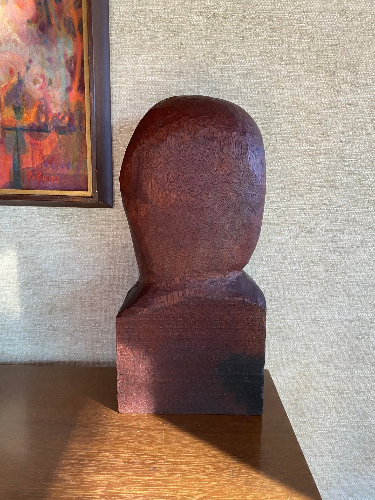 Wooden sculpture of a figure against a textured wall with a painting in the corner.