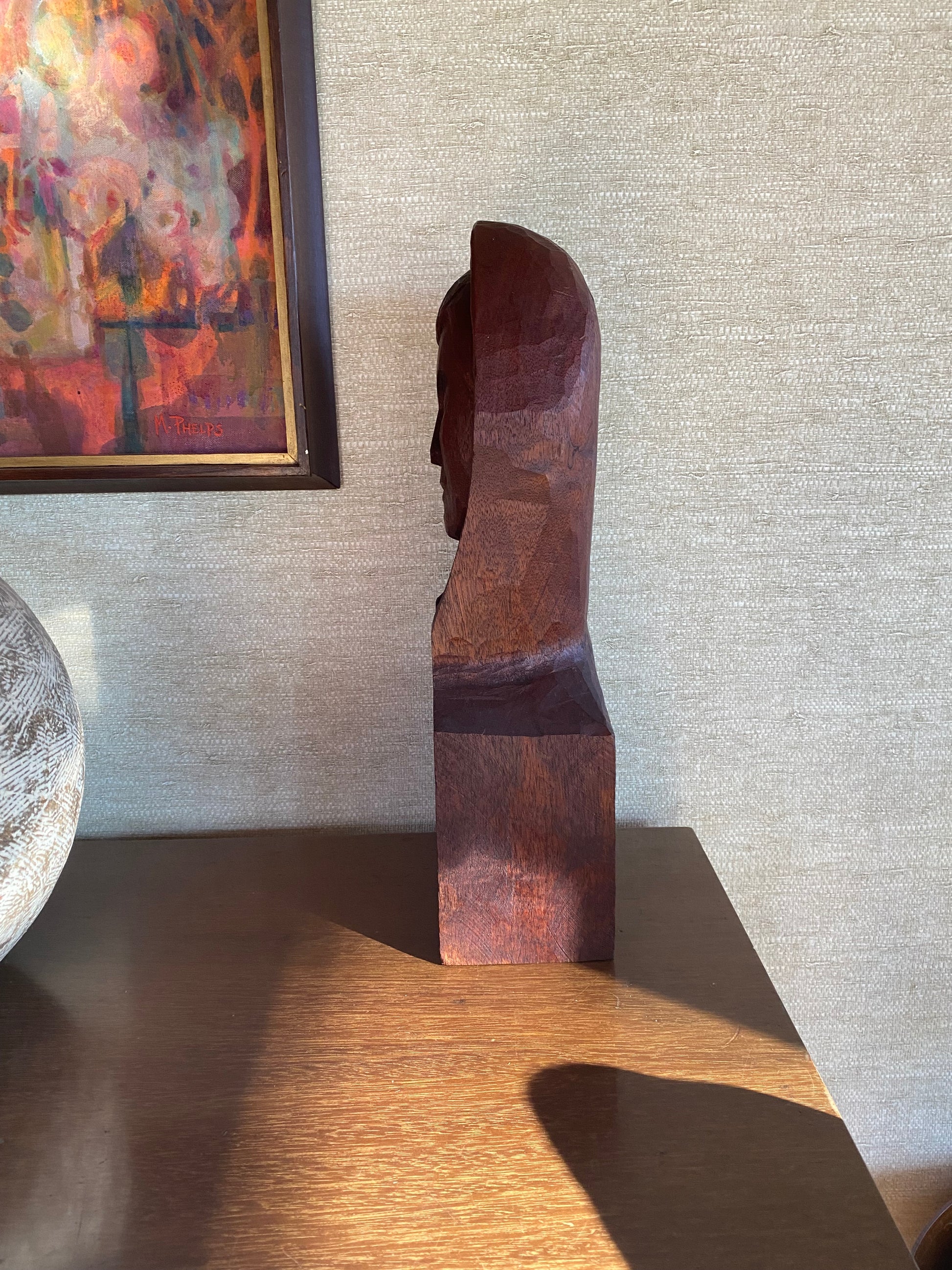Wooden bust sculpture against a textured wall with a colorful painting and decorative ball in the foreground.